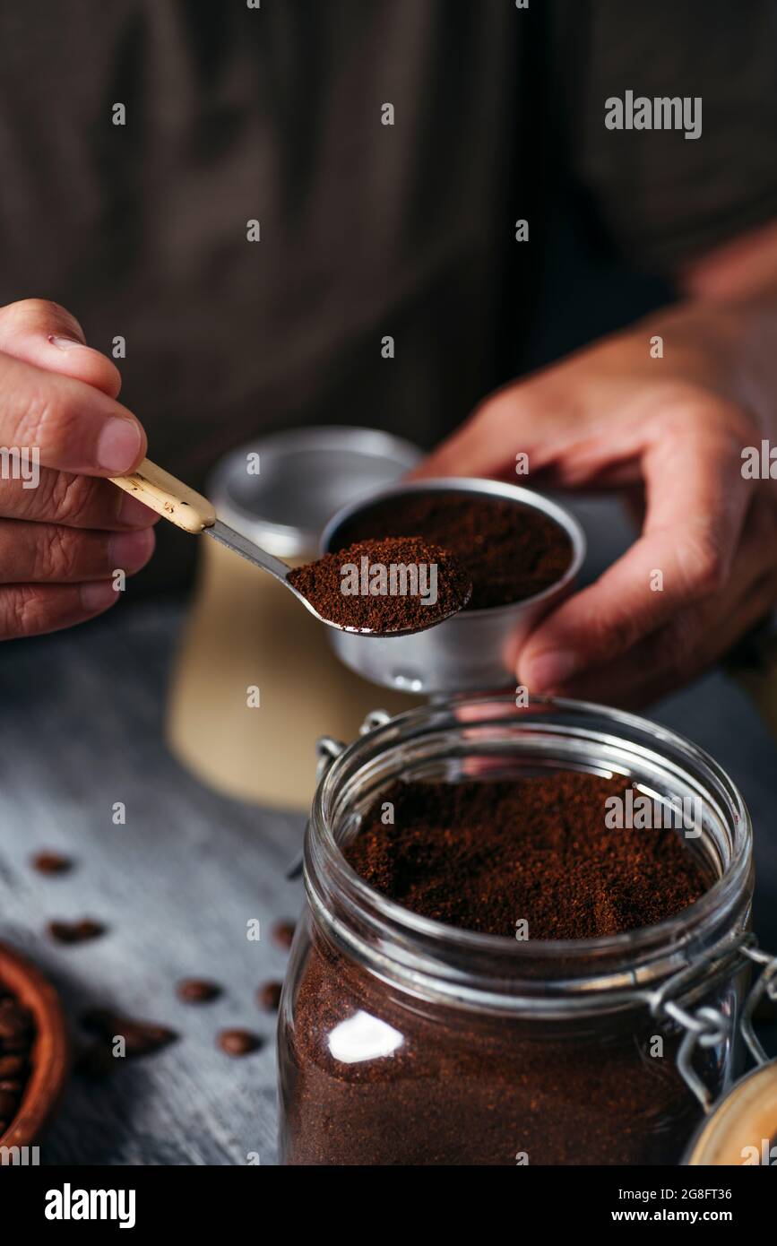 primo piano di un giovane caucasico che riempie l'imbuto di una pentola di moka con caffè macinato, da un vaso di vetro, seduto su un tavolo di legno rustico grigio Foto Stock