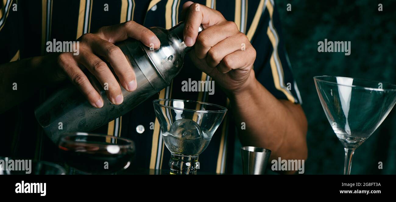 un giovane caucasico, indossando una camicia a righe, prepara un cocktail in uno shaker metallico, in un formato panoramico da utilizzare come banner web o intestazione Foto Stock