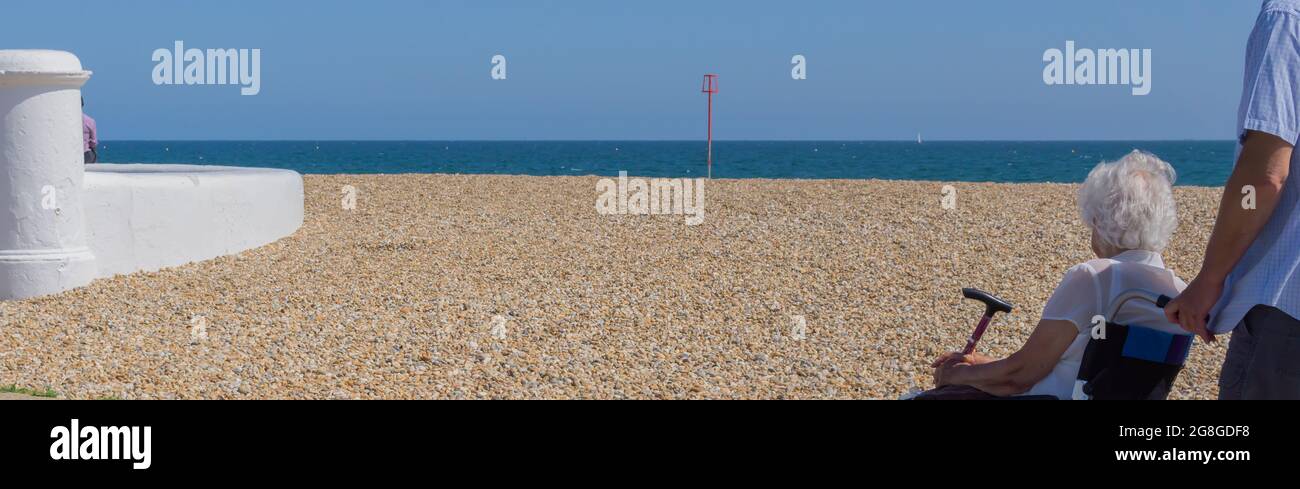 Una vista laterale e posteriore di un anziano sconosciuto in una sedia a rotelle con assistente, guardando verso una spiaggia Pebble e vista mare. Foto Stock
