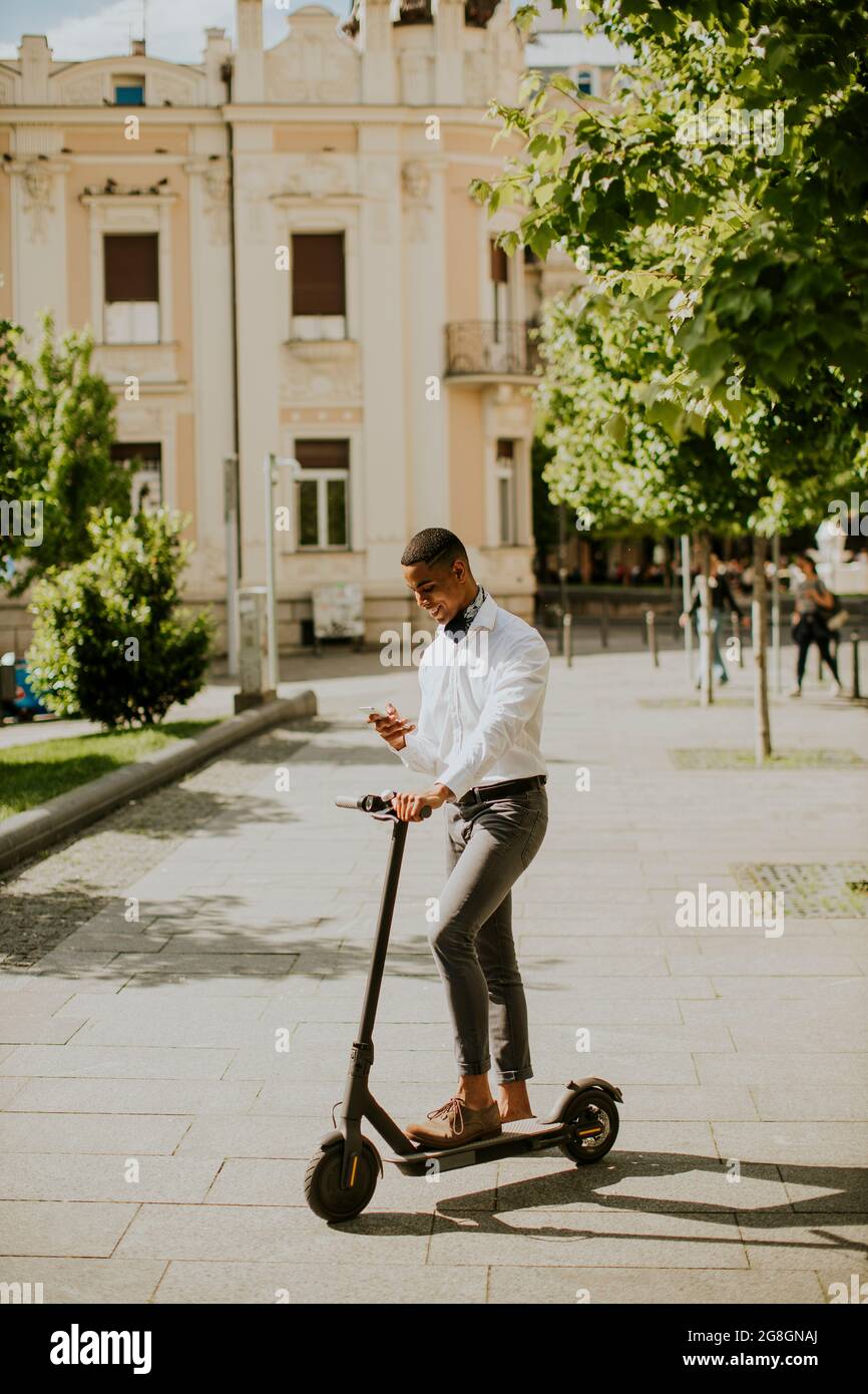 Bel giovane afroamericano che usa il telefono cellulare mentre si è in piedi con uno scooter elettrico su una strada Foto Stock