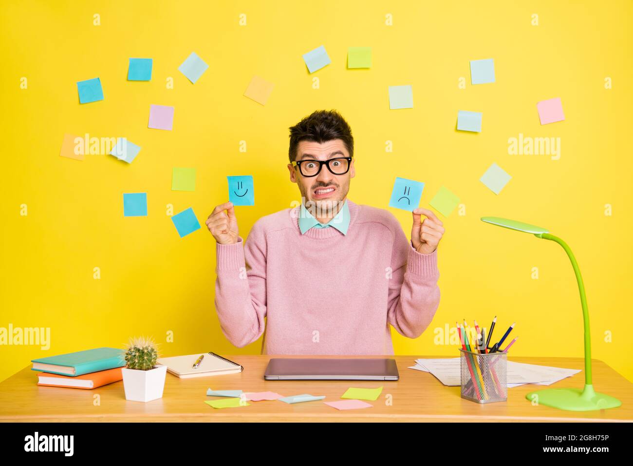 Foto di giovane infelice turbato uomo triste preoccupato in occhiali mostrando emoji di carta felice e triste isolato su sfondo giallo Foto Stock