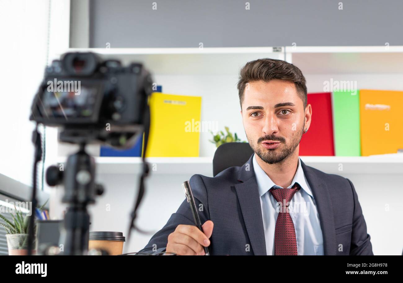 Uomo d'affari sicuro seduto alla scrivania e guardando la fotocamera sul treppiede. Giovane uomo in tuta che usa la trasmissione in diretta in streaming via internet. Foto Stock