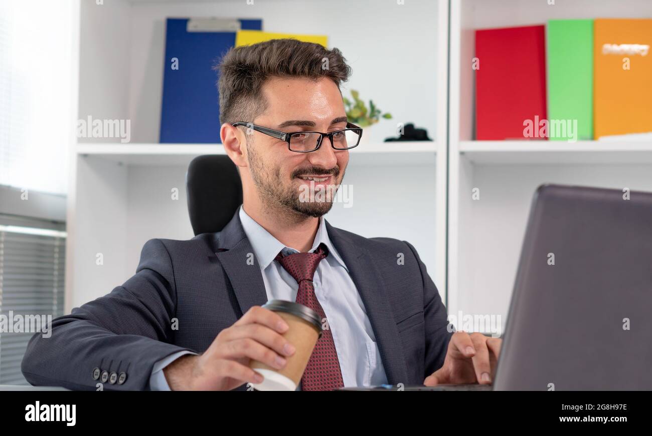 Ritratto di felice uomo d'affari in abbigliamento formale che lavora con il computer portatile in ufficio. Uomo allegro e rilassato in tuta che lavora mentre beve caffè o tè. Foto Stock