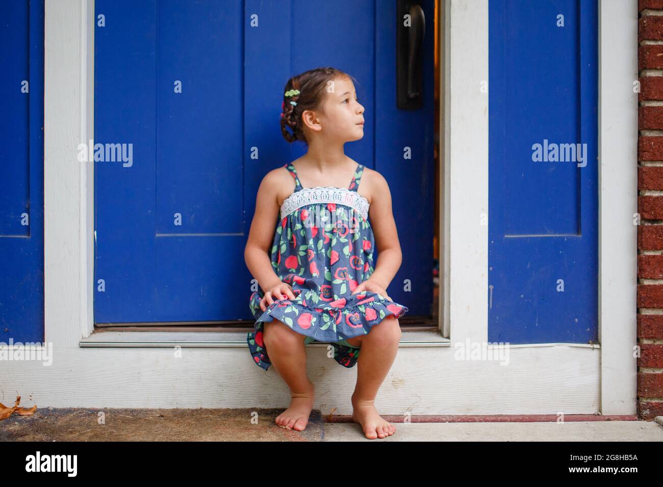 Carino ragazza piccola con trecce siede a piedi nudi in Sundress sul fronte stoop Foto Stock