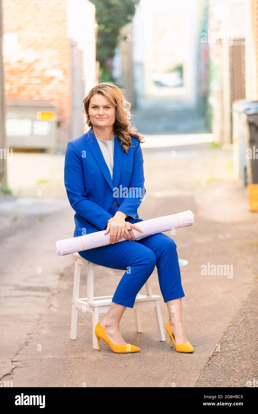 Corpo pieno di elegante donna d'affari di mezza età in abito lucido blu  pantaloni e scarpe gialle che tengono tirato e guardando la fotocamera  mentre si siede su u Foto stock -