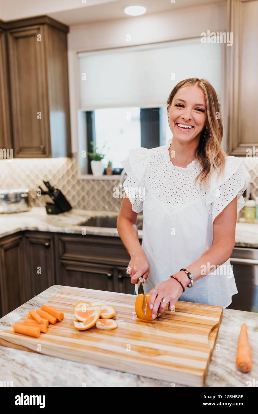 Donna sorridente affetta un arancio su un tagliere nella sua cucina Foto Stock