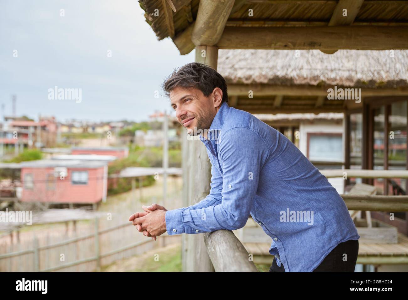Giovane uomo in una camicia blu chiaro appoggiato sulla ringhiera di legno della terrazza della sua cabina mentre sorride. Foto Stock