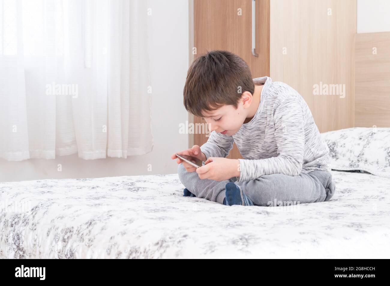 Bambino di 6 anni che usa e gioca su smartphone. Simpatico ragazzo che gioca con lo smartphone a letto. Boy gioca in videogioco sul suo smartphone. Foto Stock