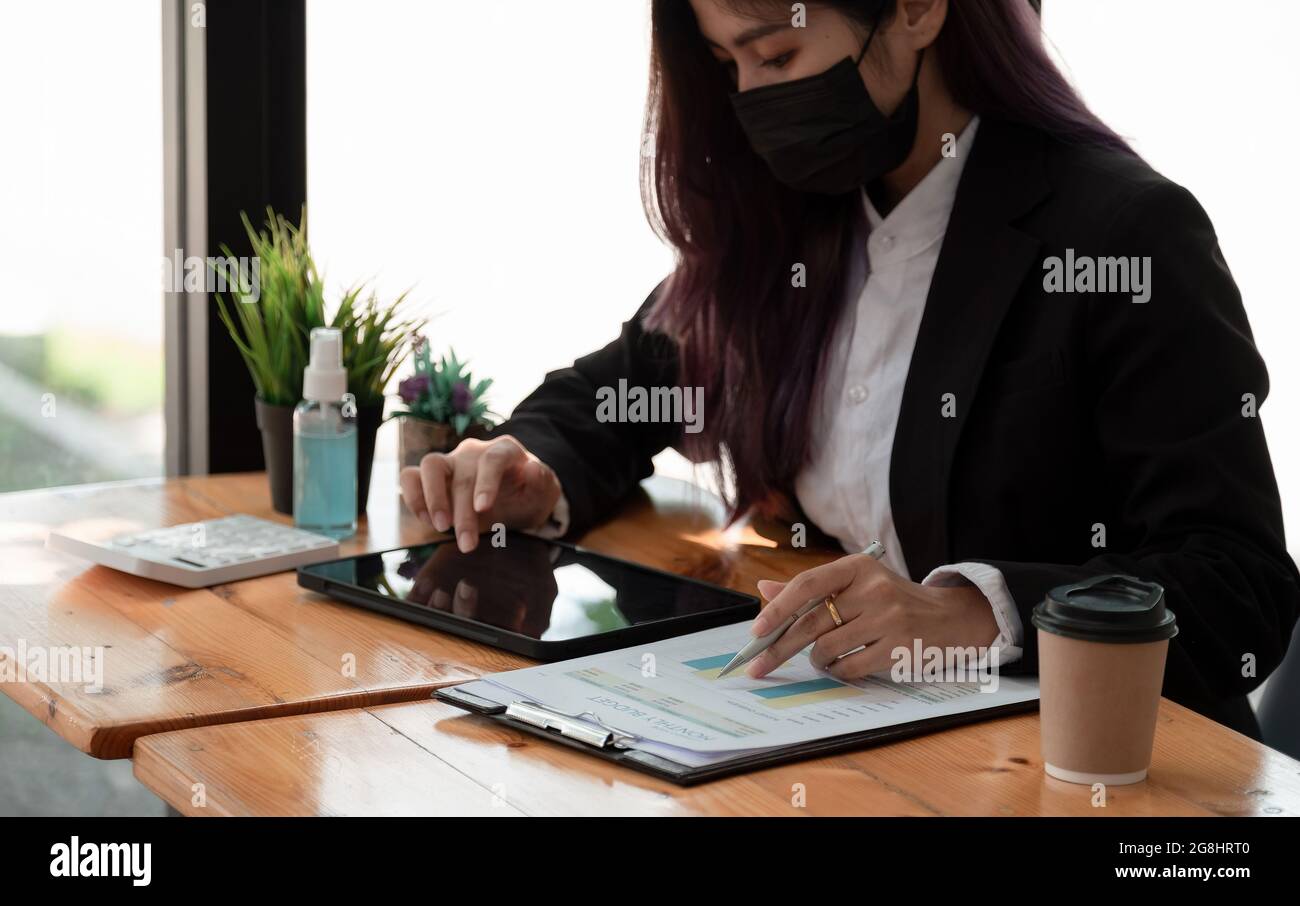 Vista laterale di una donna d'affari asiatica che indossa una maschera mentre si lavora su un computer per i calcoli finanziari in ufficio. Concetto di igiene e sicurezza. Foto Stock