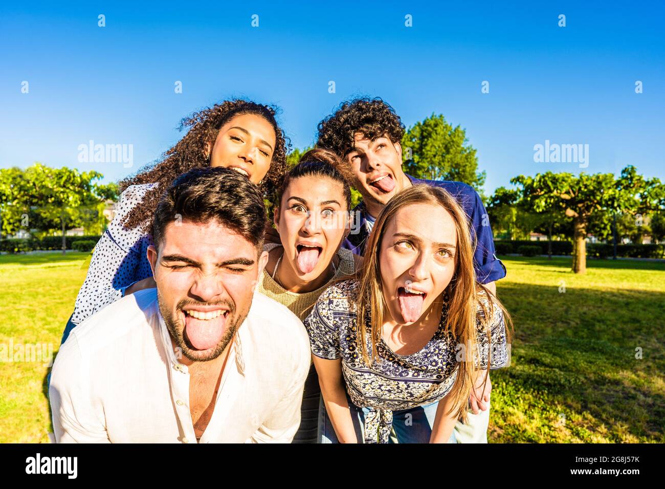 Gruppo di giovani sciocchi multirazziali amici millenari che fanno volti divertenti con la lingua, la bocca aperta, e squinting occhi in posa per un ritratto nel parco della città. Foto Stock