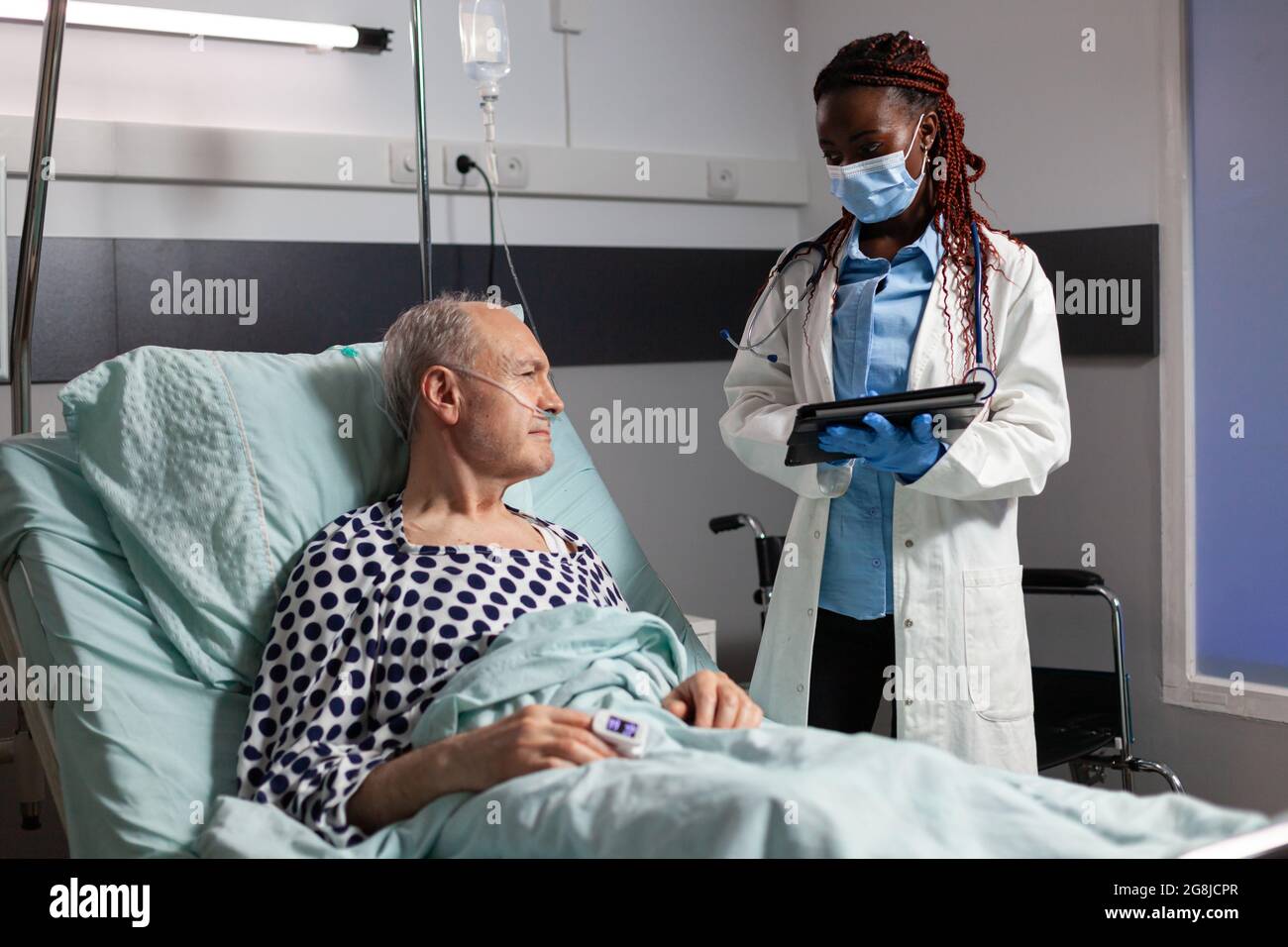 Medico afroamericano con maschera chirurgica in sala d'ospedale che discute diagnosi e trattamento con l'uomo anziano malato che si posa a letto rassicurando il recupero completo. Paziente malato che respira attraverso la maschera di ossigeno. Foto Stock