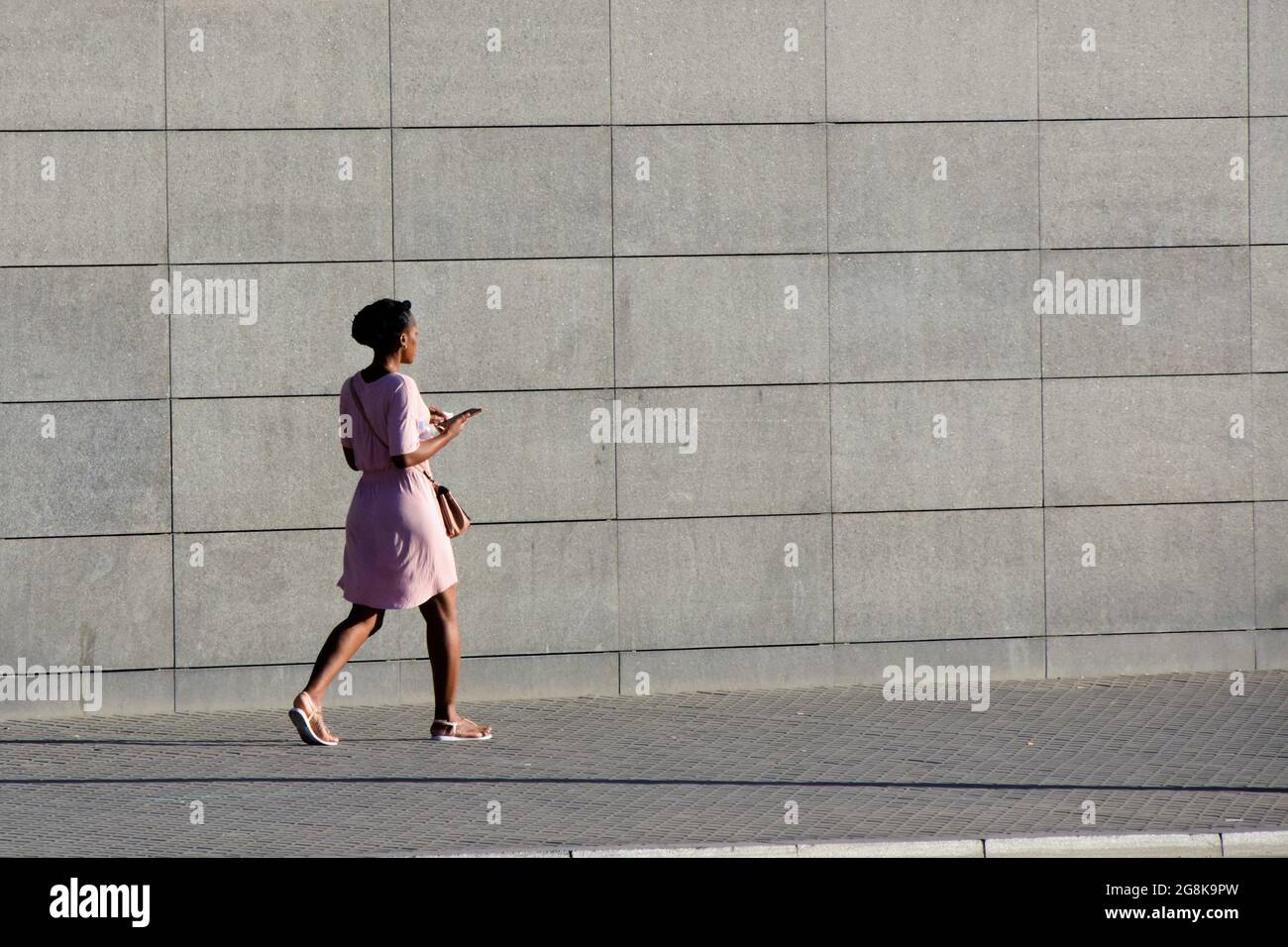 Giovane donna nera sta camminando vicino a un muro grigio in abiti estivi mentre sta tenendo il suo smartphone. Foto Stock