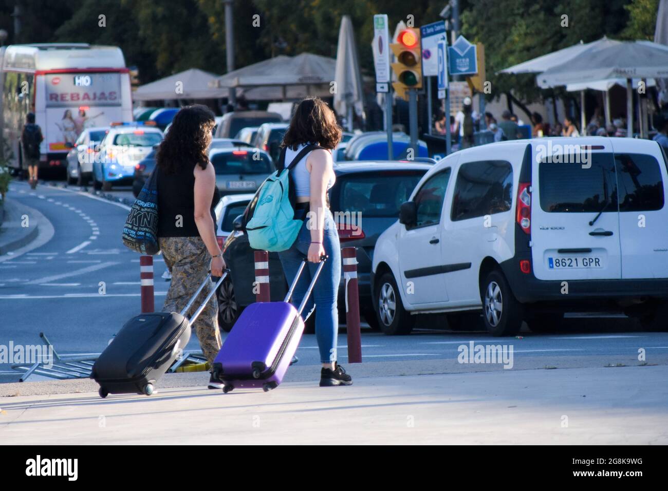 Due giovani donne che camminano per strada con le valigie e gli zaini gommati in estate. Vista del traffico, coda di auto in attesa al semaforo rosso. Foto Stock