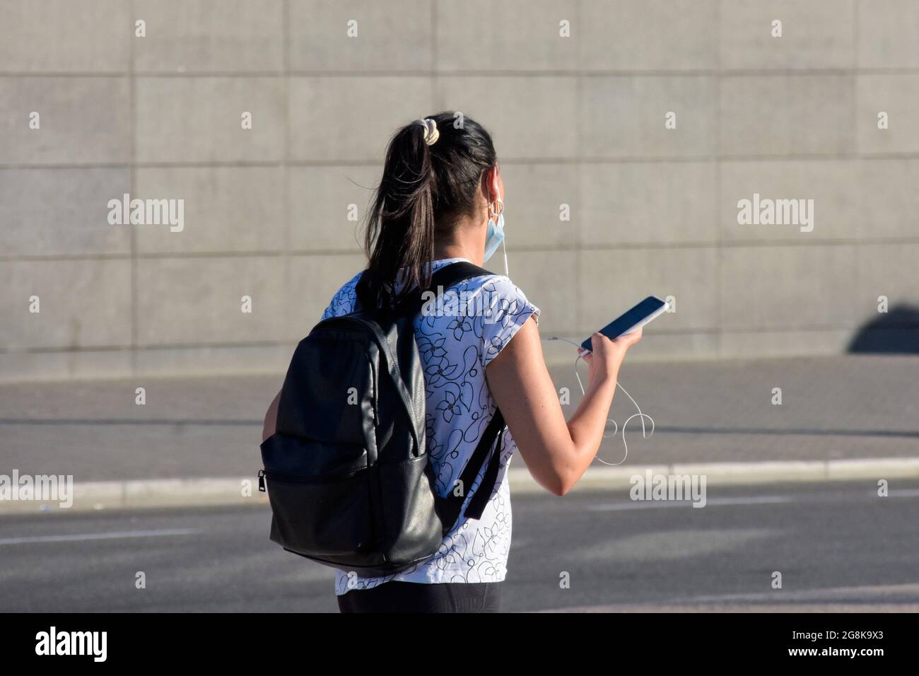 Vista posteriore di una giovane donna che tiene uno smartphone e ascolta con gli auricolari mentre cammina in una strada. Foto Stock