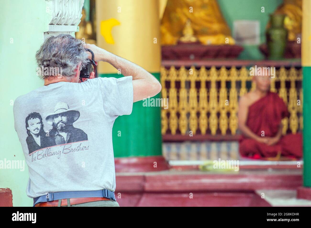 Western tourist indossare Bellamy Brothers t-shirt scatta foto di vecchio monaco (fuori fuoco) seduto in posizione di loto nel Tempio di Shwedagon, Yangon, Myanmar Foto Stock