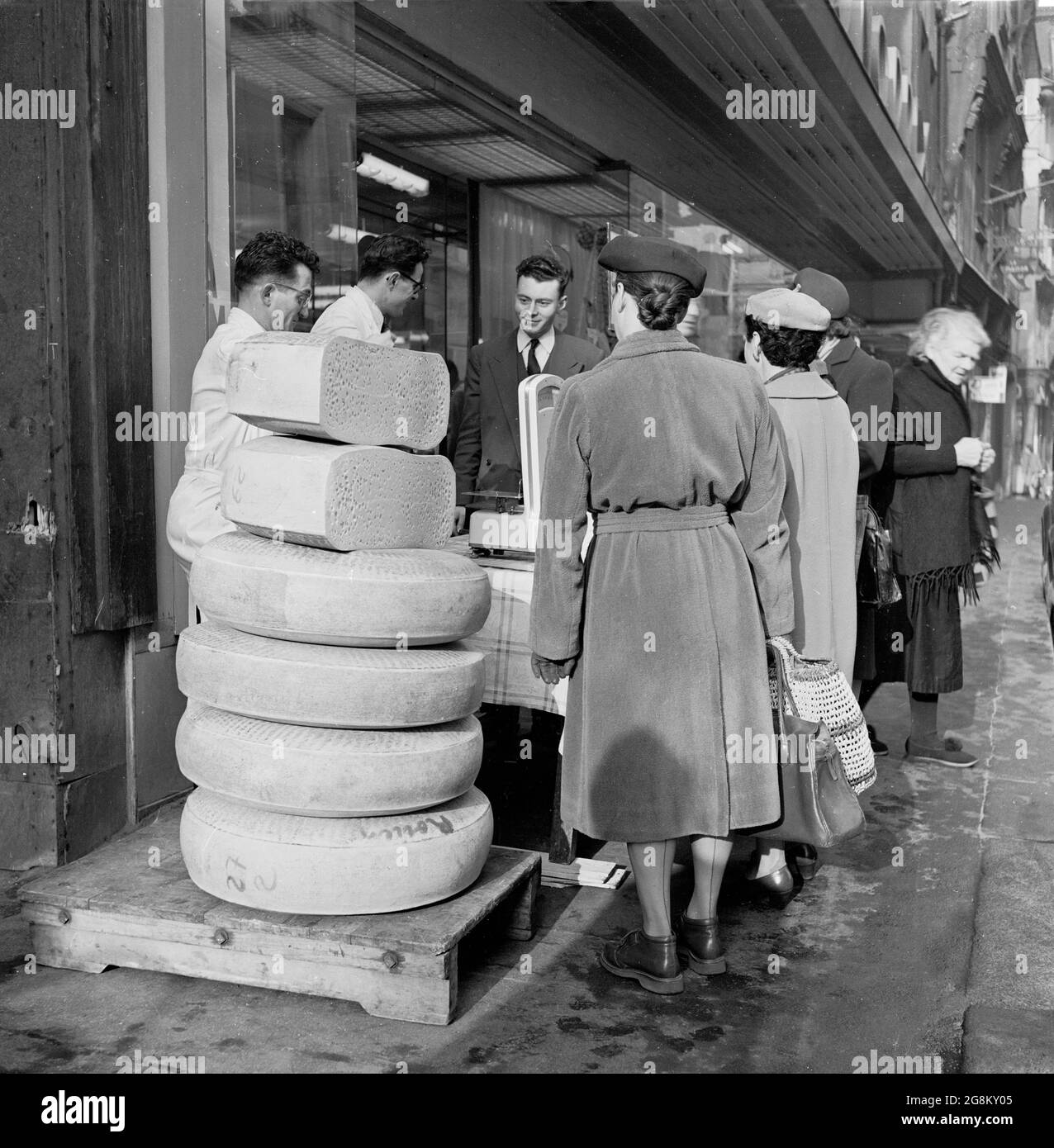 Immagine storica che mostra le donne che fanno shopping a Rouen all'esterno della strada, in piedi da alcune grandi fette rotonde di formaggio accatastate su una piattaforma di legno all'esterno di un negozio di formaggi o une Cremerie, Francia anni '50 Foto Stock