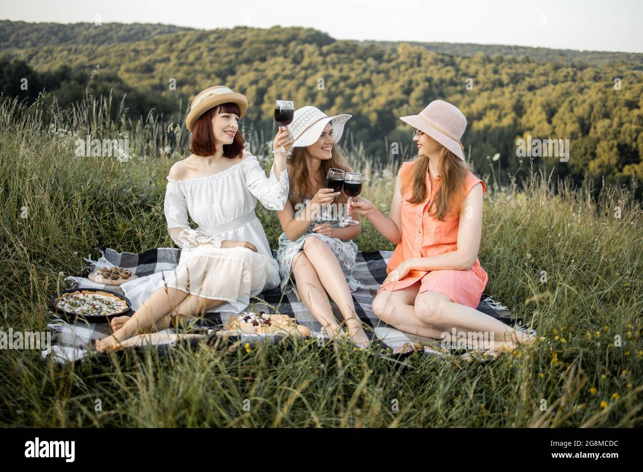 Gruppo di tre giovani donne sbalorditive in un pic-nic nel campo.  Sorridendo i migliori amici che bevono vino rosso sul prato. Ragazze in  abiti e cappelli eleganti che si rilassano e pranzano