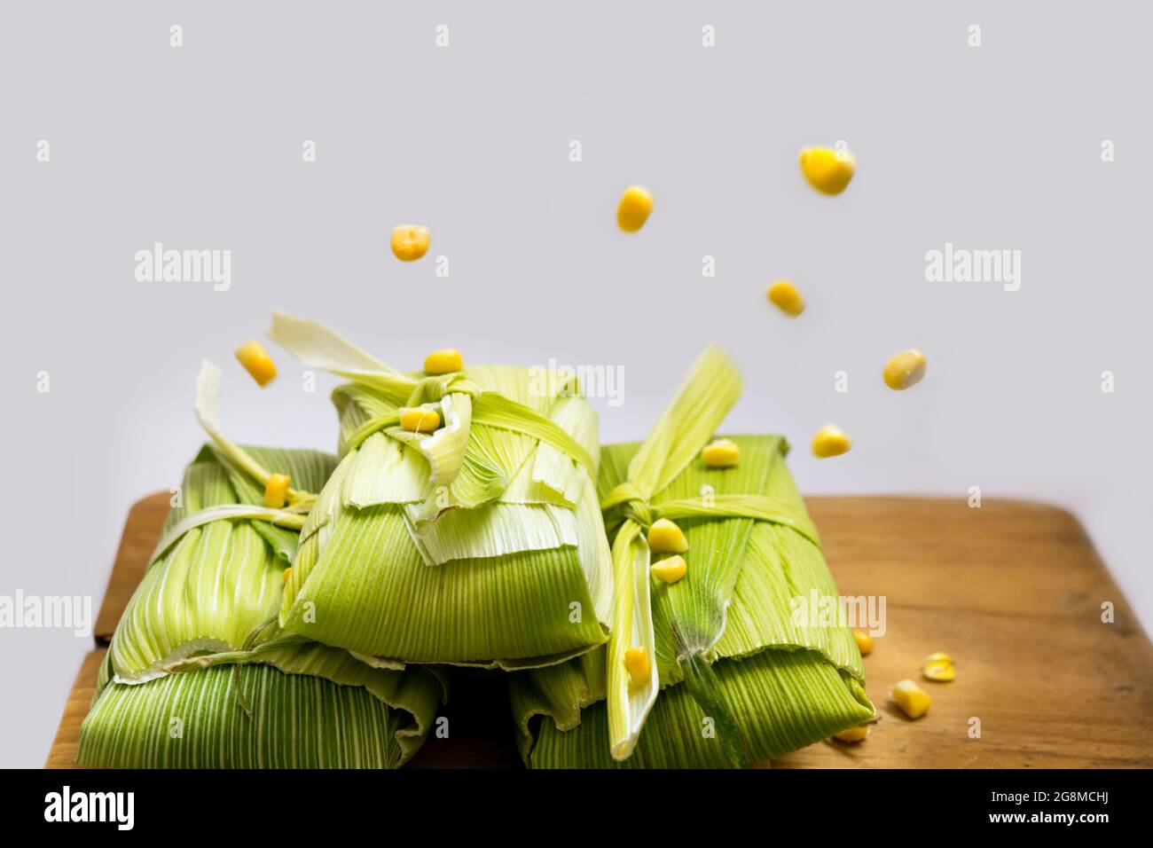 Primo piano di tre pacchetti di tamale su un tavolo di legno e alcuni grani grezzi di mais nell'aria che cade sui pacchetti, alcuni già sui pacchetti Foto Stock