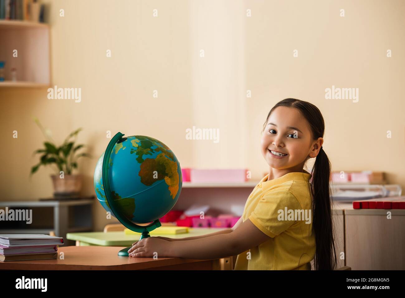 gioiosa ragazza asiatica sorridente a macchina fotografica vicino a globo nella scuola di montessori Foto Stock