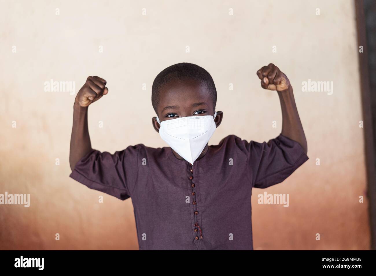 Questo è un ritratto di un bambino africano con le braccia piegate per mostrare forza. Indossa una maschera protettiva per evitare la diffusione della Th Foto Stock
