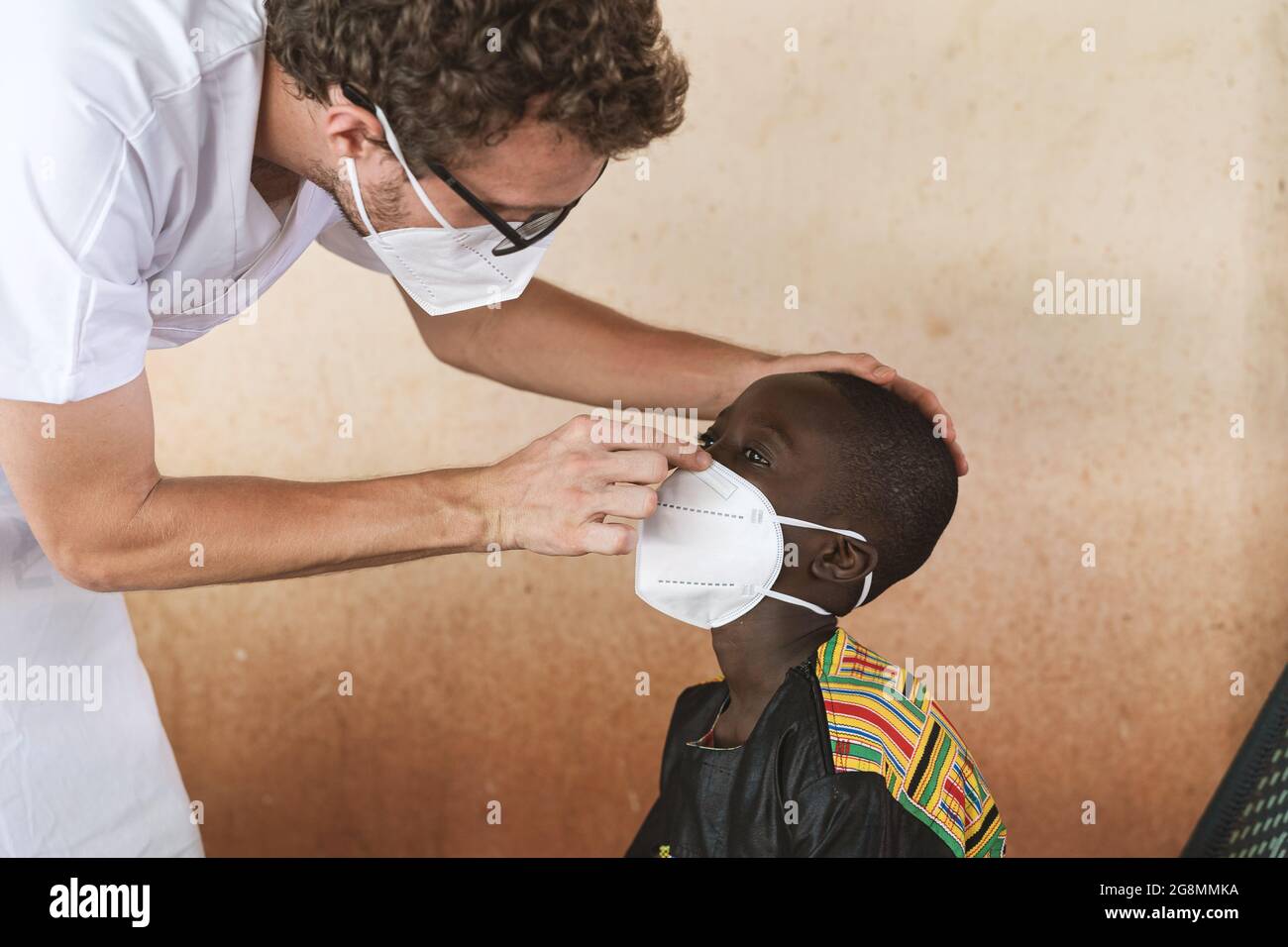 Questa è un'immagine di un medico di volontariato caucasico che aiuta un bambino africano nero a mettere correttamente la sua maschera protettiva sul viso. Foto Stock