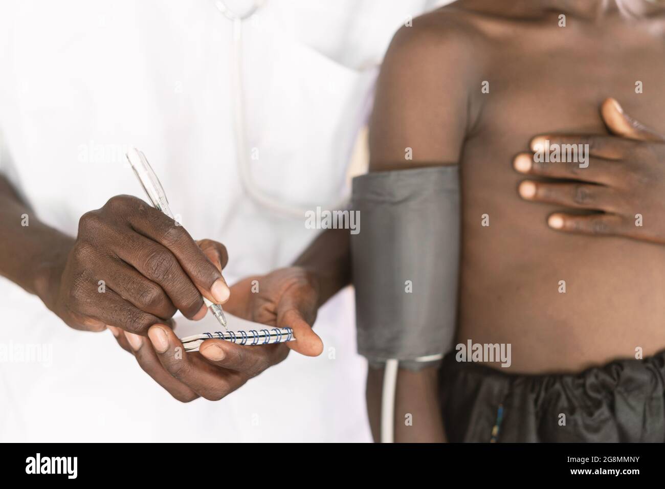 Questa immagine mostra un dispositivo di misurazione della pressione sanguigna sul braccio superiore destro di un piccolo bambino africano skinny con la mano sul petto in piedi accanto a. Foto Stock
