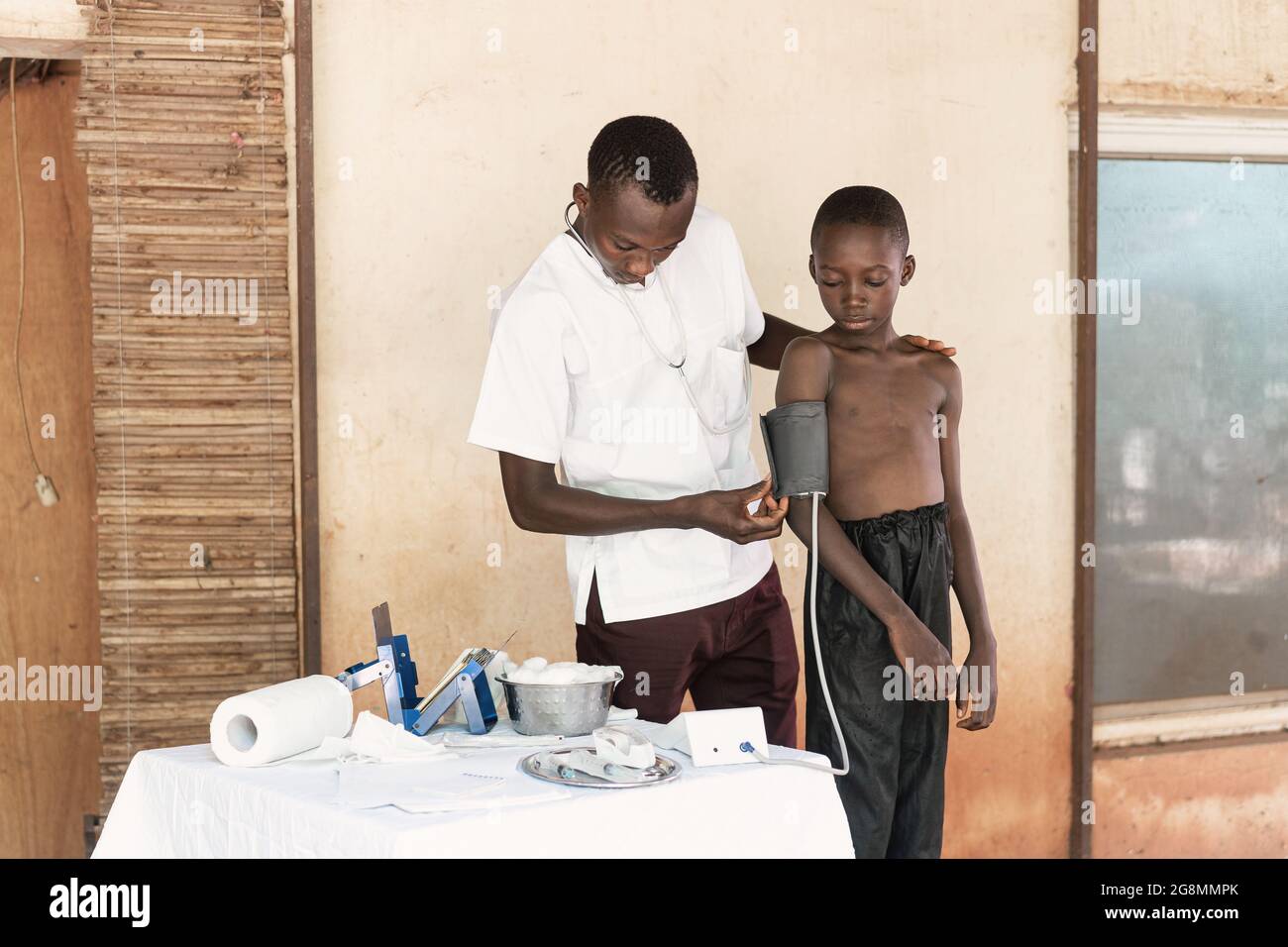 In questa immagine, un giovane operatore sanitario prende i valori della pressione sanguigna da un piccolo ragazzo nero durante il check-up di routine in un ambiente ospedaliero rurale in AF Foto Stock