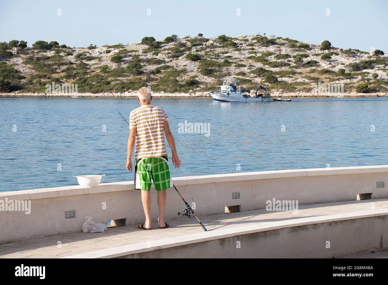 Tribunj, Croazia - 8 luglio 2021: Uomo anziano che pesca su una riva mentre pesca barca a strascico che naviga vicino Foto Stock