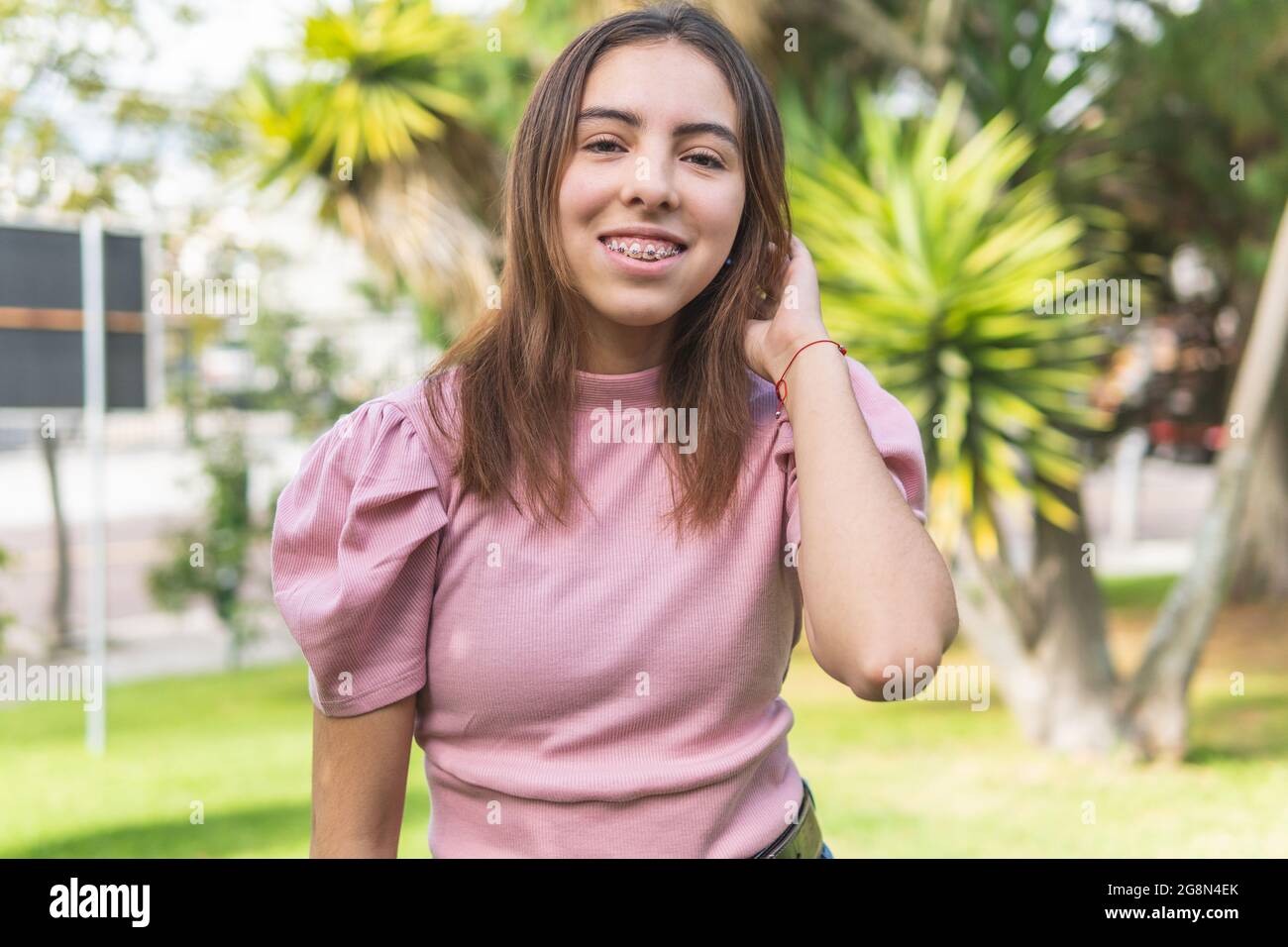 Ritratto verticale di una teenager Latina con bretelle sorridenti in un parco e stuzzicante i suoi capelli lisci leggeri Foto Stock