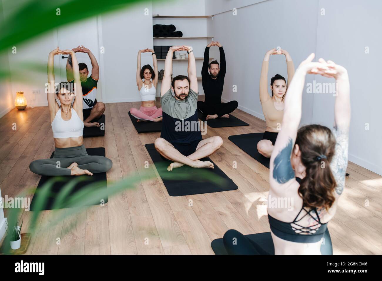 Persone che lavorano sodo che praticano lo yoga in un gruppo, facendo allungamenti in posa facile in un grande studio. Davanti a uno strumentore, ragazza con tatuaggio alle spalle. Foto Stock