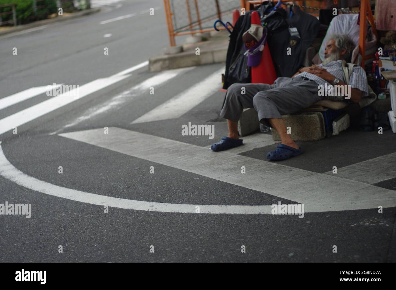 Uomo senza tetto che dorme nella striscia mediana con i suoi vestiti e altri beni di famiglia sotto la strada della città terra ponte elevato, in Giappone, giorno nuvoloso Foto Stock