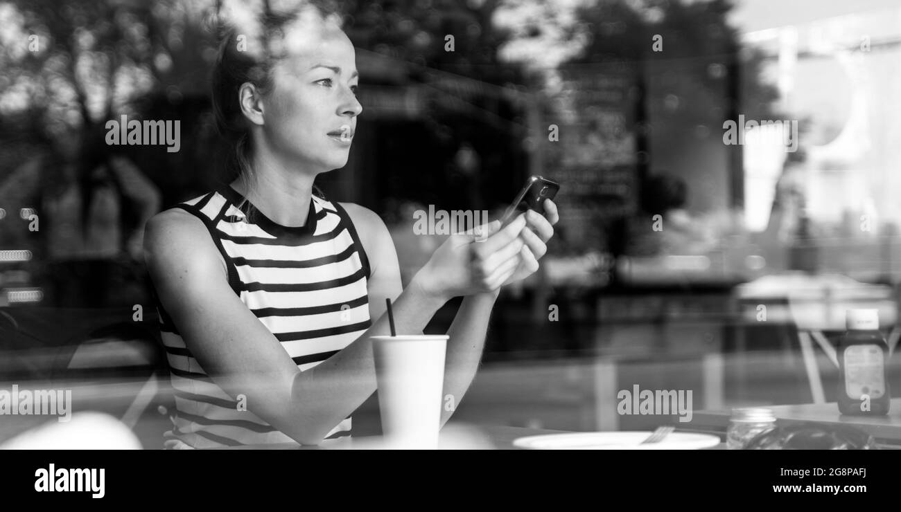 Premurosa donna caucasica tenendo il telefono cellulare mentre si cerca attraverso la caffetteria finestra durante la pausa caffè. Foto Stock