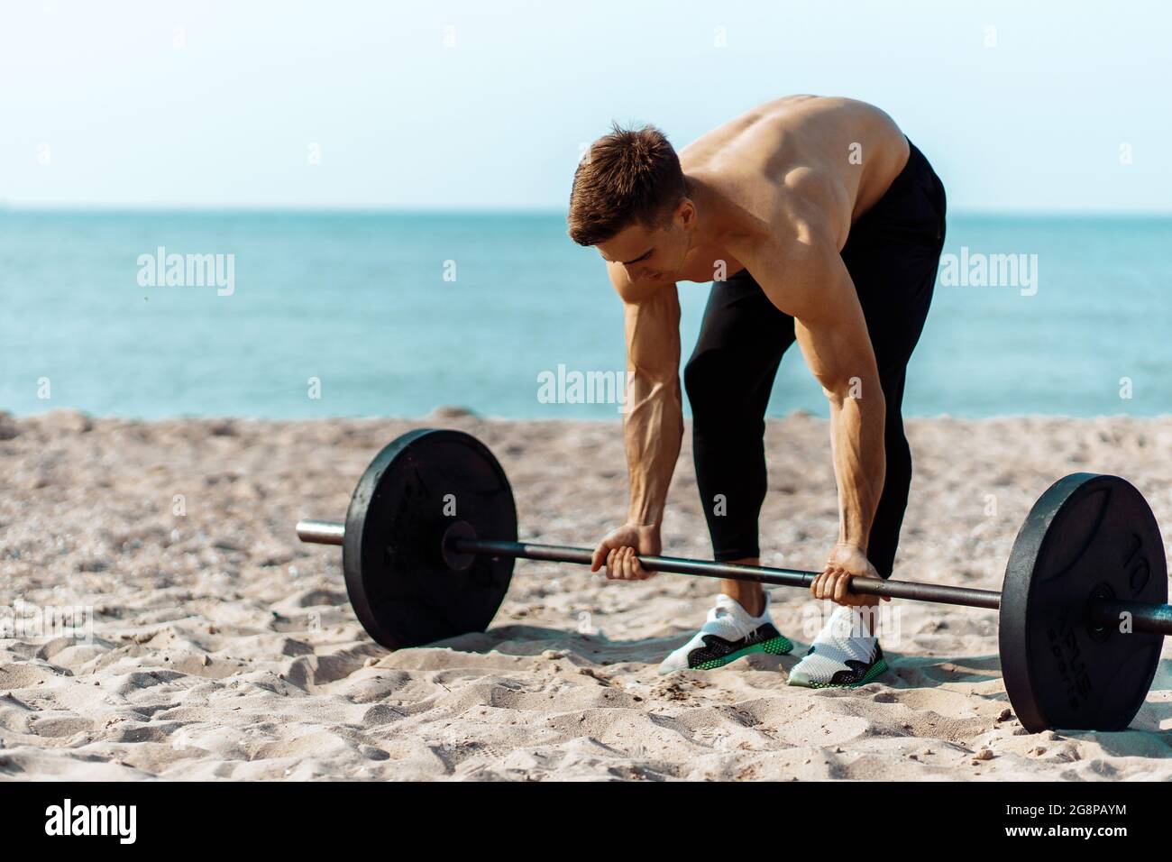 Muscular Guy bodybuilder fare esercizi con grandi manubri all'aperto sulla spiaggia, in una giornata estiva di sole, allenarsi sulla costa del mare sullo sfondo Foto Stock