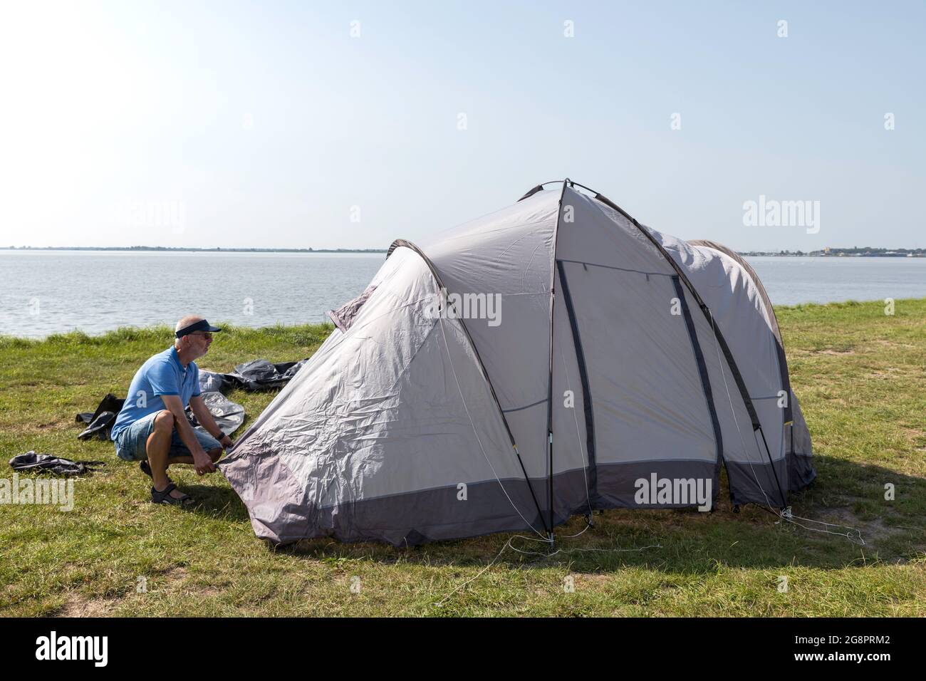 uomo maturo che stabilisce una tenda e che mette giù i picchetti della tenda su un campo vicino all'acqua su uno spazio bello vicino al lago Foto Stock