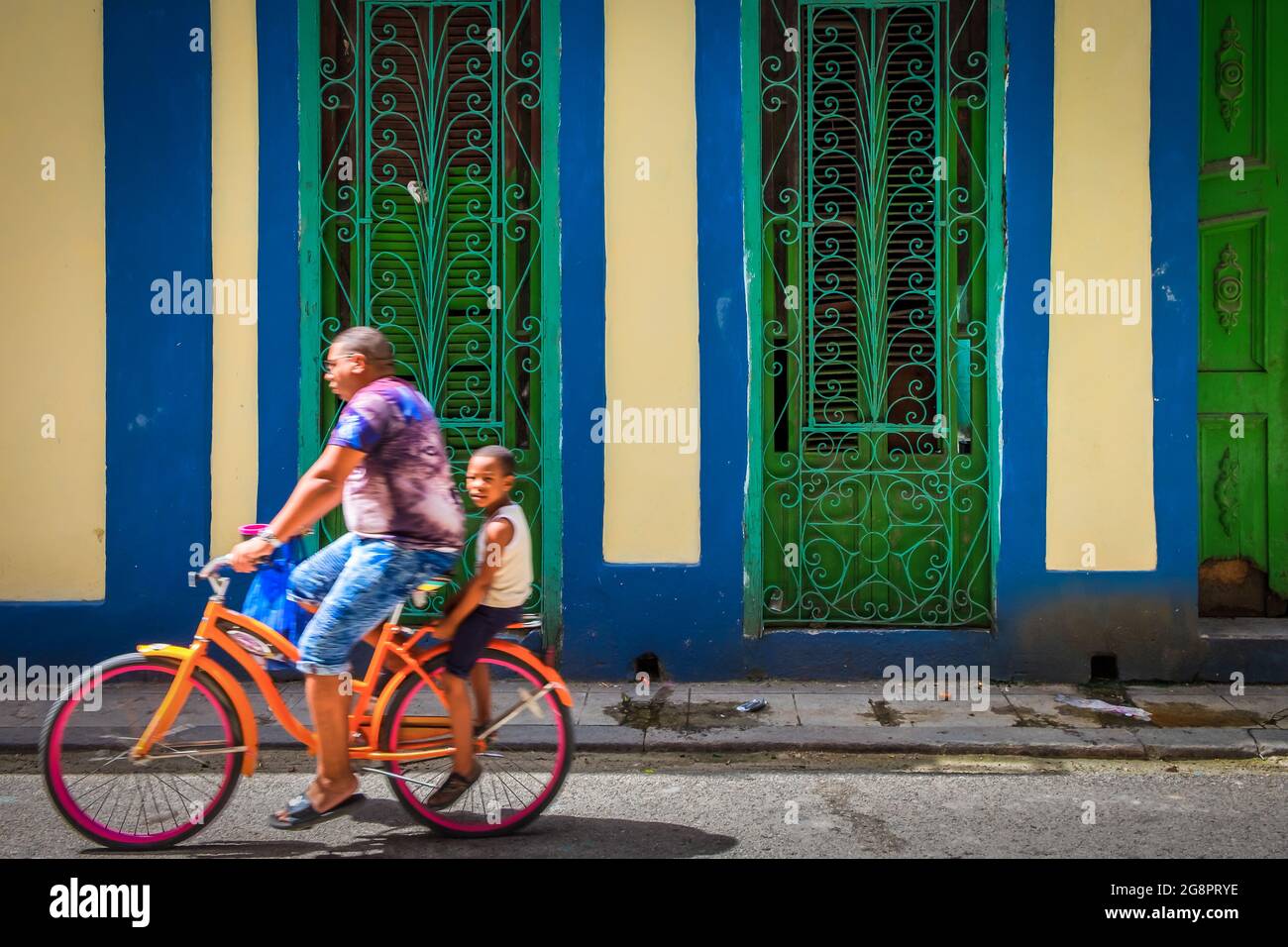 Havana, Cuba, 2019 luglio, un padre e un figlio che cavalcano una bicicletta arancione accanto a un colorato muro della casa nella parte vecchia della città Foto Stock