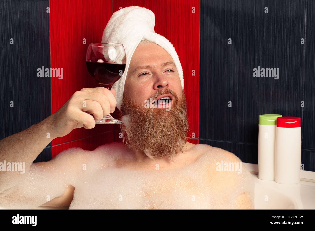 Felice uomo bearded che prende bagno di bolla. Divertente ragazzo in turbante capelli rilassarsi con un bicchiere di vino rosso in una vasca da bagno schiumosa e godersi la vita dopo una giornata difficile. Foto Stock