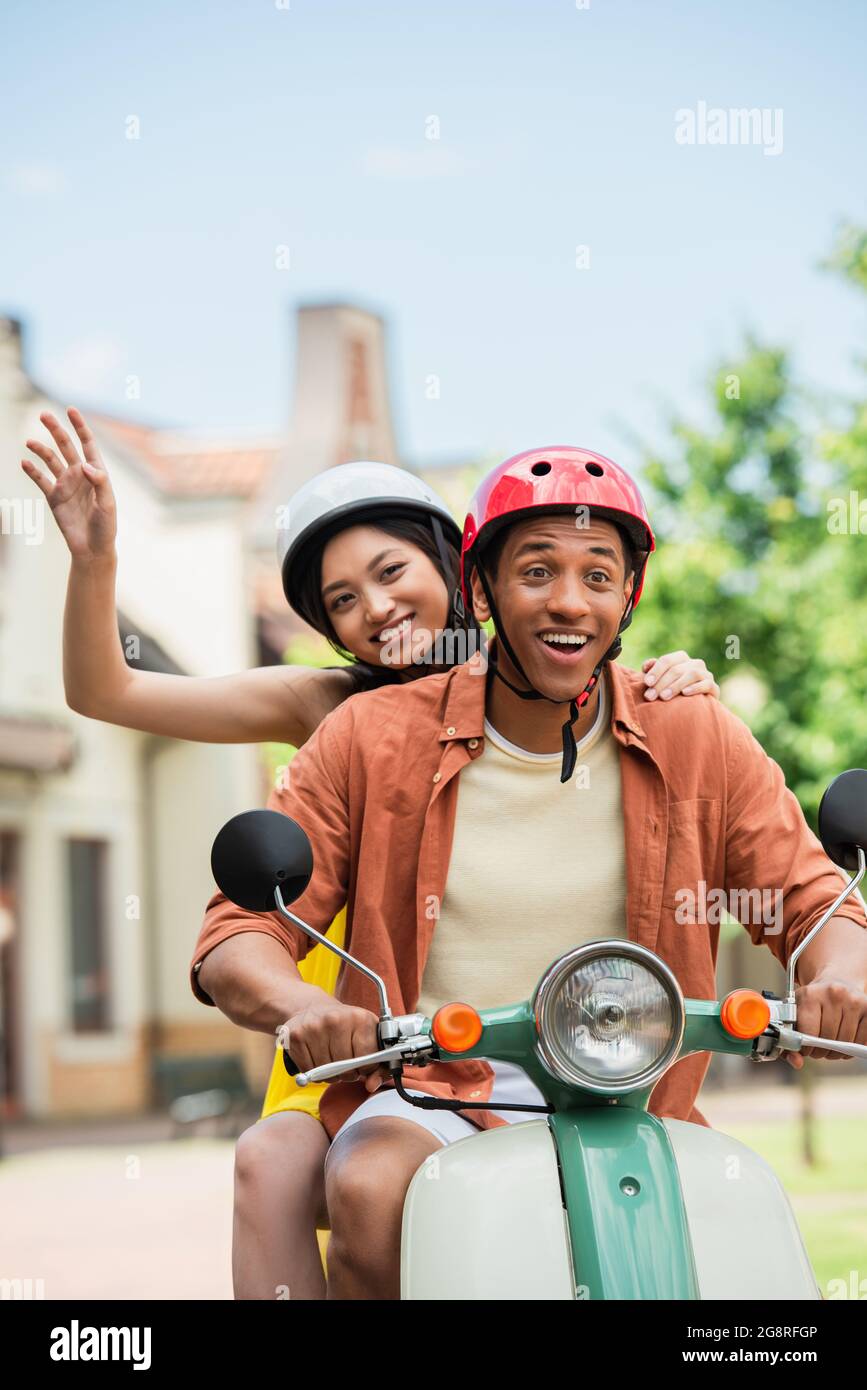felice donna asiatica che svetta mano mentre si guida scooter con l'entusiasta uomo ameriano africano Foto Stock
