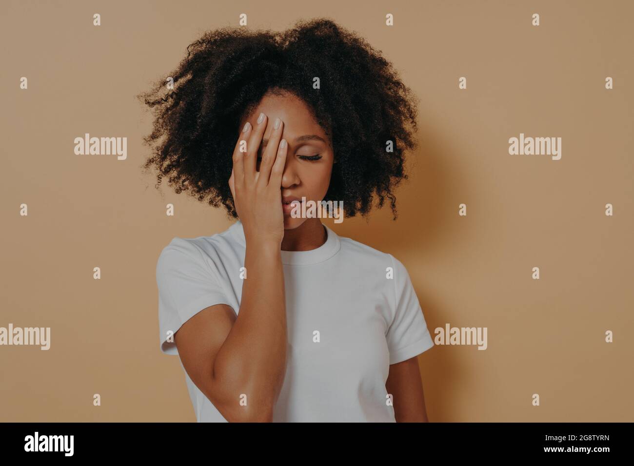 Pelle scura femmina che ricopre la faccia con la mano e sensazione di ansia o depressione contro la parete beige Foto Stock
