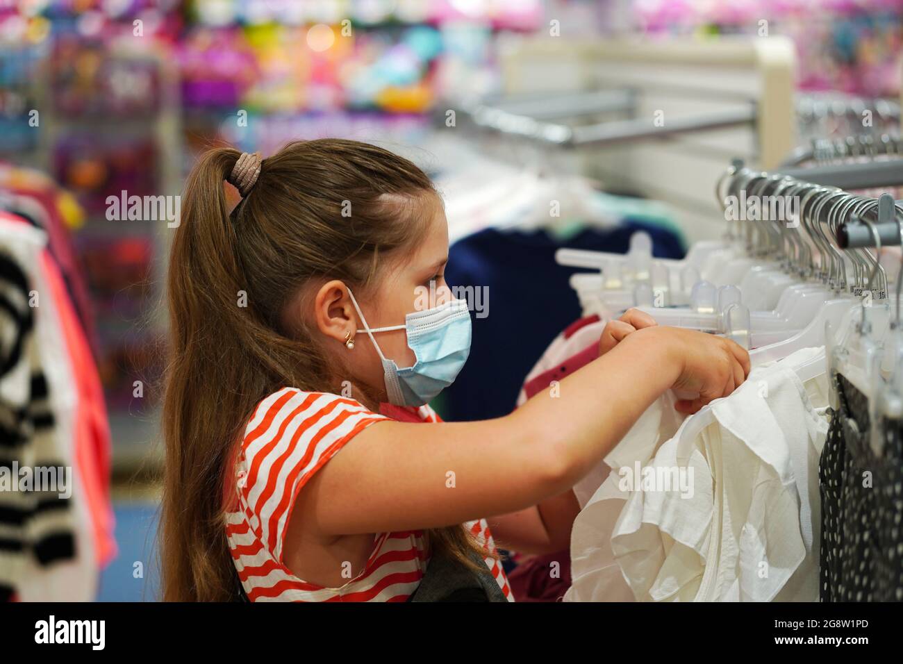 La bambina in una maschera medica protettiva sceglie il deposito uniforme della scuola. Preparazione per la scuola. Prevenzione del coronavirus. Torna a fare shopping a scuola. Foto Stock