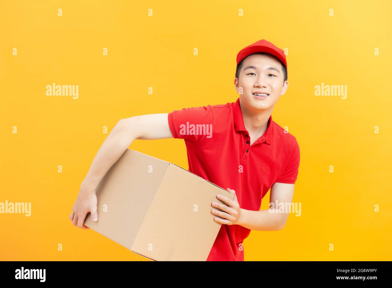 felice giovane addetto alla consegna in piedi con tappo rosso con scatola posta isolata su sfondo giallo Foto Stock