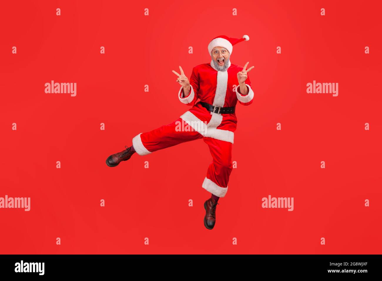 Ritratto a lunghezza intera di uomo anziano felice con barba grigia che indossa il costume di babbo natale saltando mostrando v gesto con le dita, celebrando la bufera invernale Foto Stock