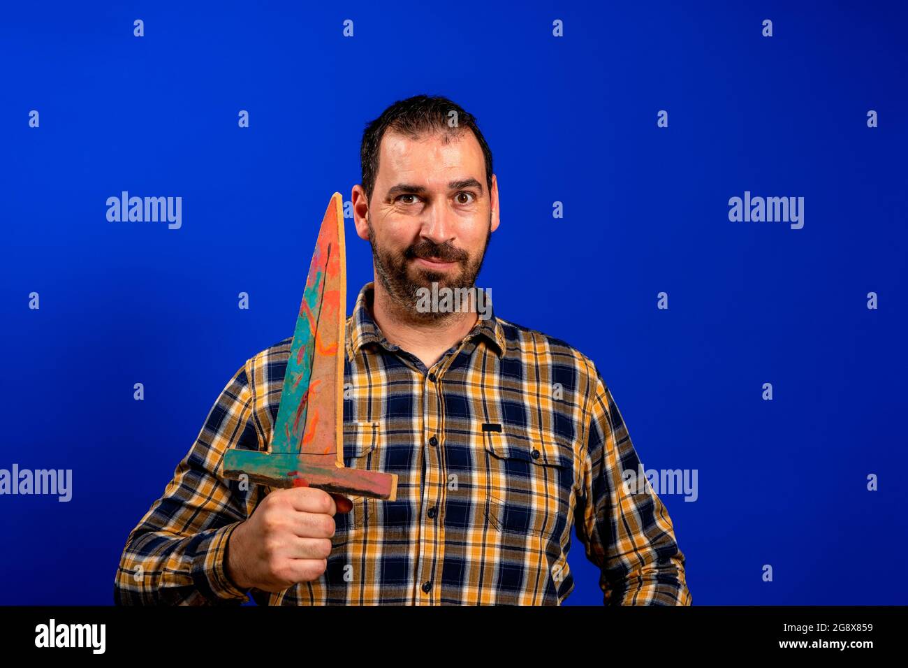 Uomo in una camicia gialla in plaid graffiando la schiena con una spada di legno isolata su sfondo blu studio. Foto Stock