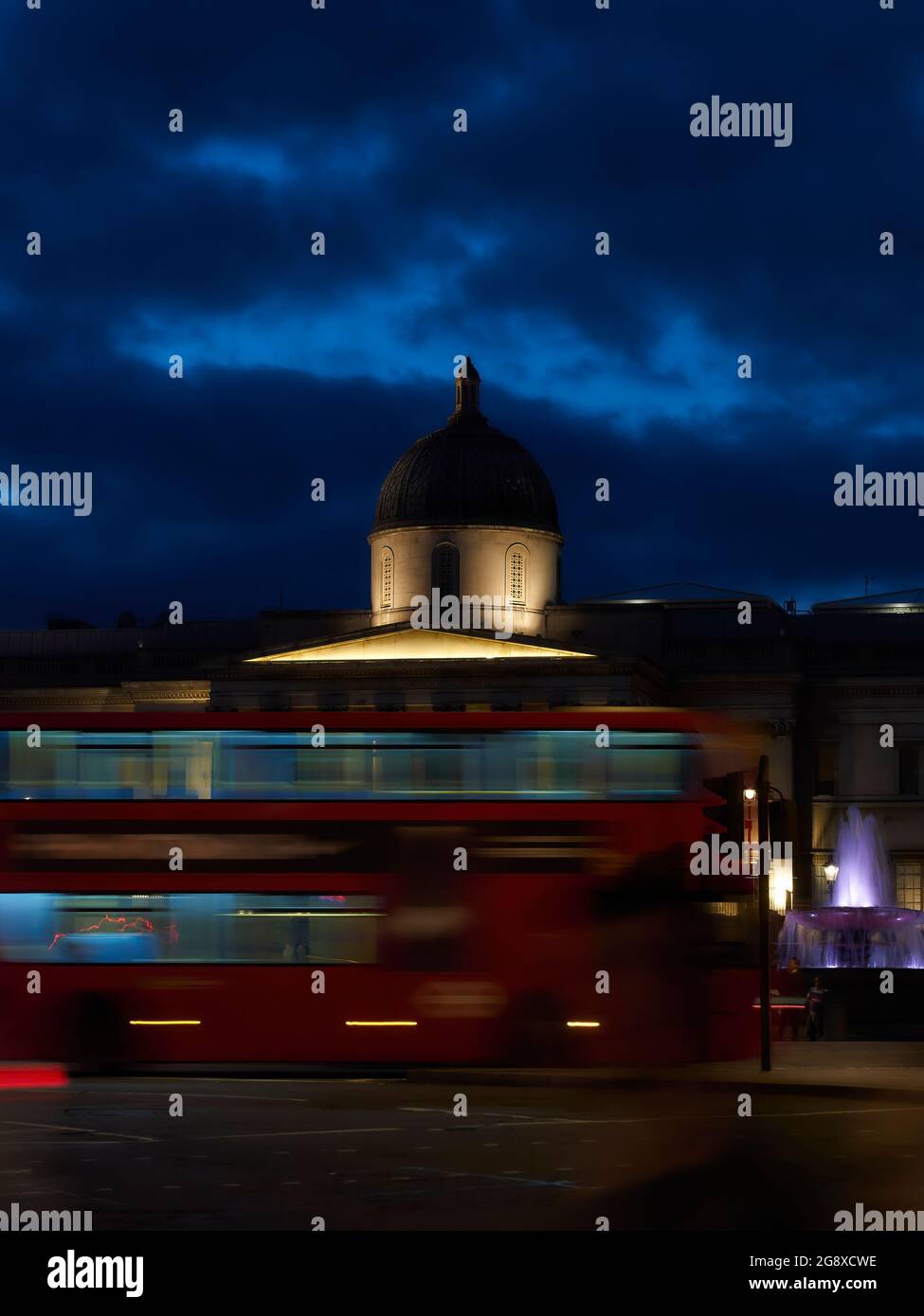 Immagine energica che mostra un autobus londinese sfocato nel movimento che passa di fronte a una National Gallery gridata di notte in Trafalgar Square. Foto Stock
