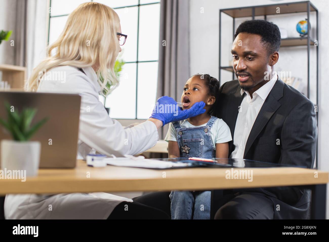 Concetto di assistenza sanitaria e pediatria. Vista posteriore del medico femminile biondo che controlla la gola della bambina afro con la spatola durante l'ispezione generale. Carina ragazza visita clinica moderna con padre premuroso Foto Stock