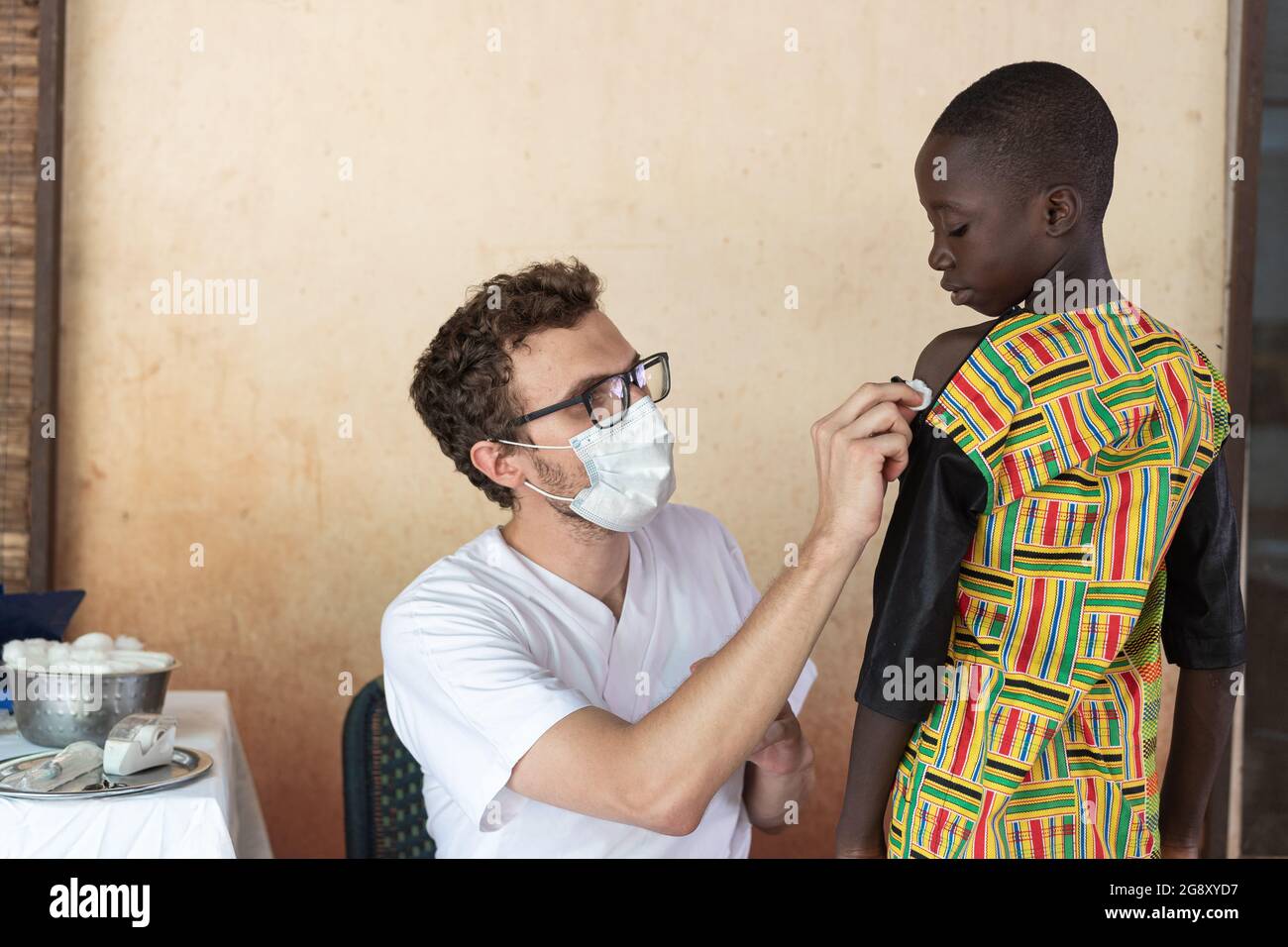 In questa immagine, un giovane medico bianco che indossa una maschera protettiva sta disinfettando il braccio di un piccolo ragazzo nero con un batuffolo di cotone prima di iniettarlo una v Foto Stock