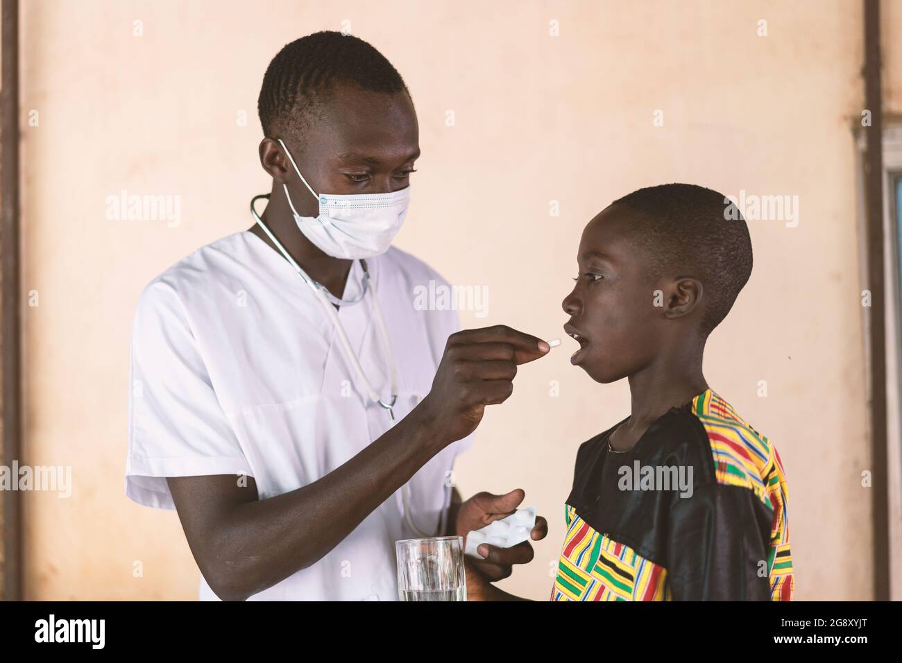 In questa immagine un giovane tirocinante medico che indossa una maschera facciale sta somministrando un medicamento orale ad un piccolo scolaro che collabora durante un'assistenza sanitaria c Foto Stock