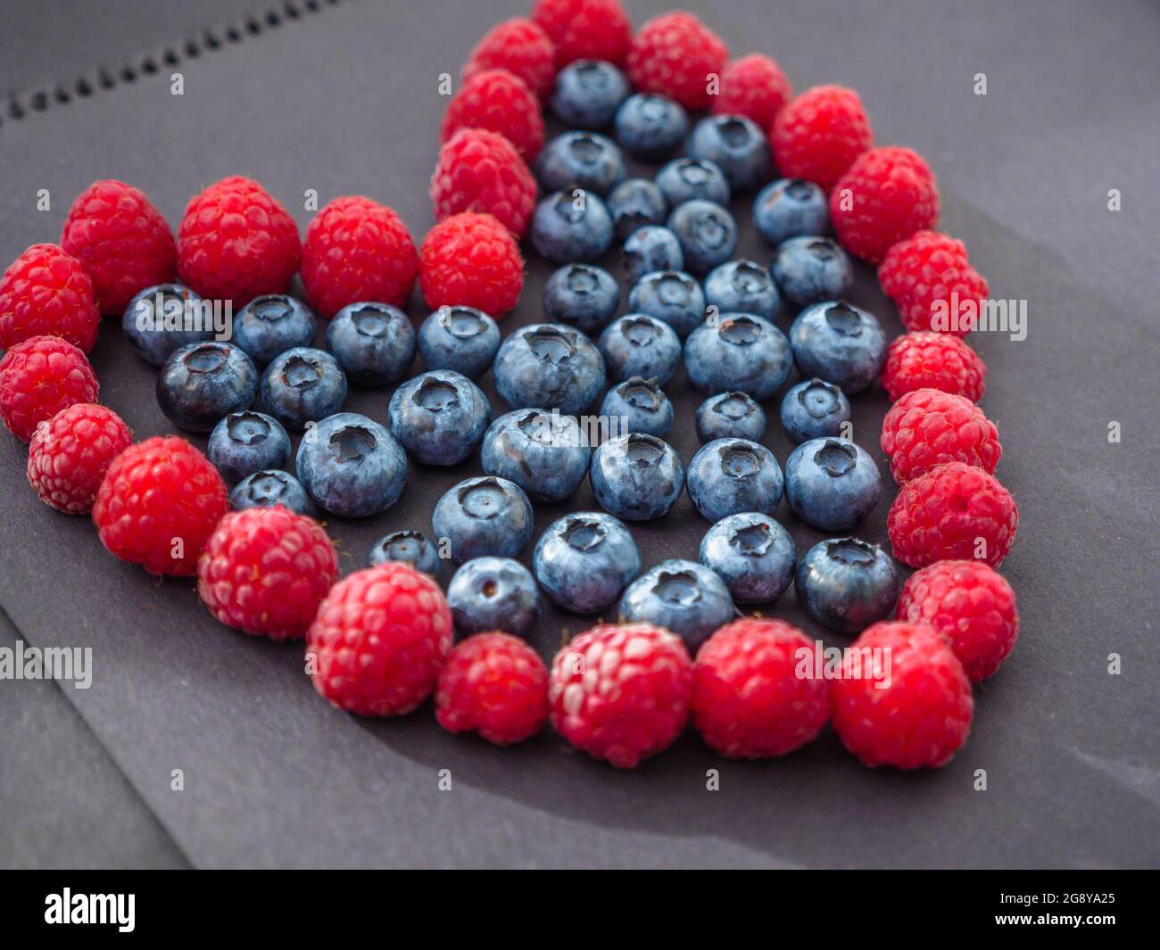 Segno d'amore a forma di cuore fatto da lamponi freschi e mirtilli su carta nera. Amore e concetto di San Valentino. Foto Stock