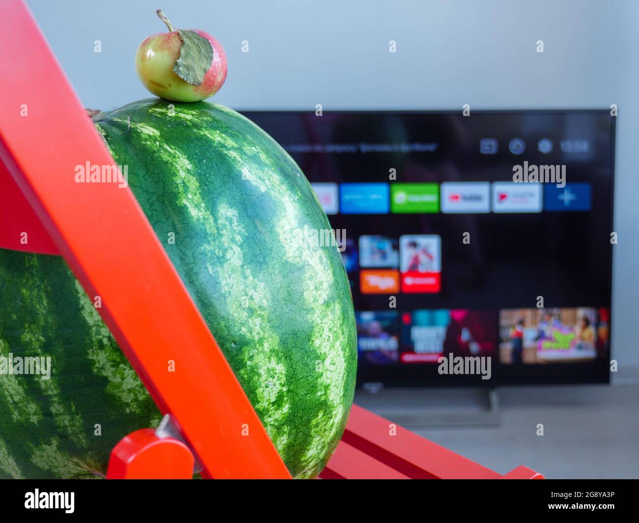 Piccola mela rossa sulla cima di un grande cocomero sedersi su un tavolo di legno rosso e guardare la TV. Gli opposti attraggono il concetto. Foto Stock