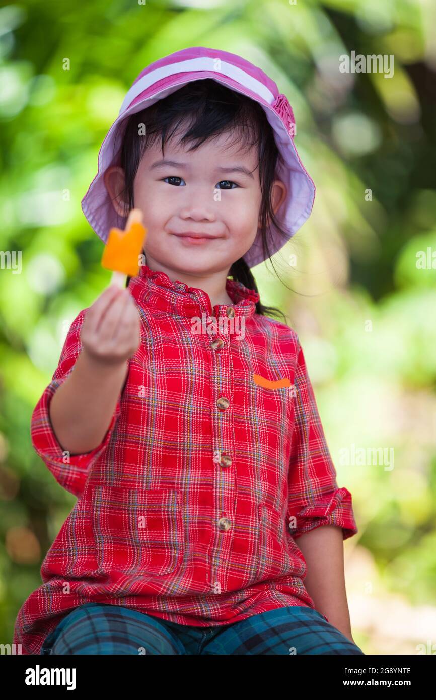 Piccola ragazza asiatica sorridente e mangiare gelato in estate su sfondo natura offuscato. Bambino grazioso in natura. All'aperto. Foto Stock