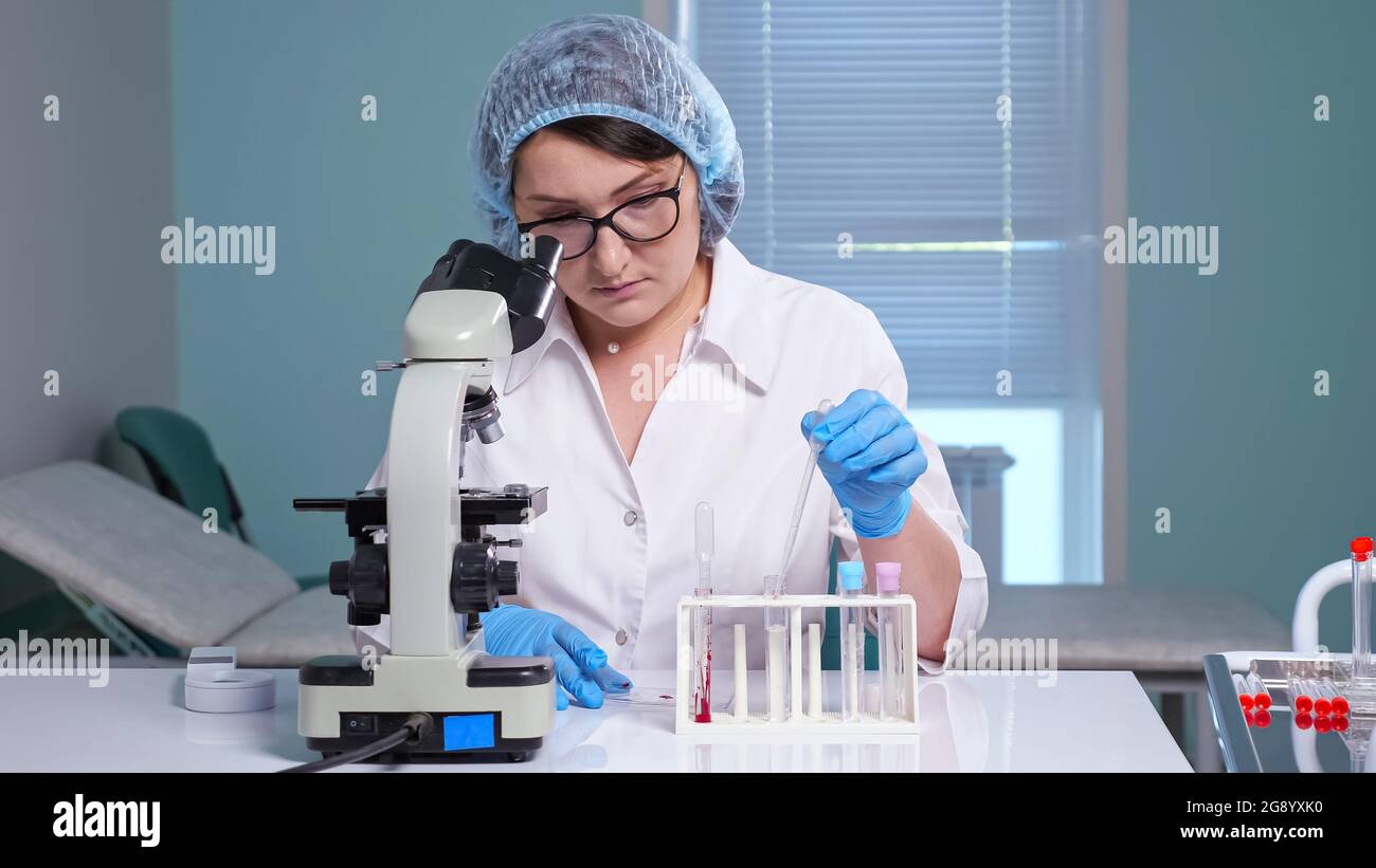L'assistente di laboratorio femminile concentrato in bicchieri e uniforme bianca preleva il liquido dal tubo e guarda al microscopio seduto al tavolo in clinica Foto Stock
